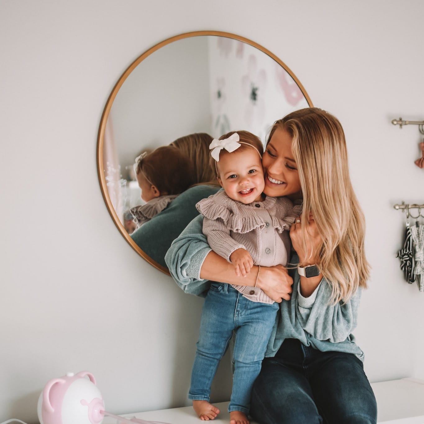 Une mère assise avec sa petite fille sur une commode à côté d'un mouche-bébé électrique Nosiboo Pro rose