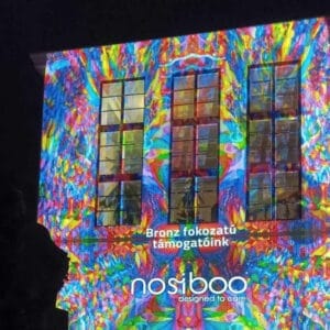 Le logo de Nosiboo s'affiche avec une lumière colorée sur un bâtiment, informant du niveau bronze du parrainage du Festival des Lumières de Zsolnay à Pécs.