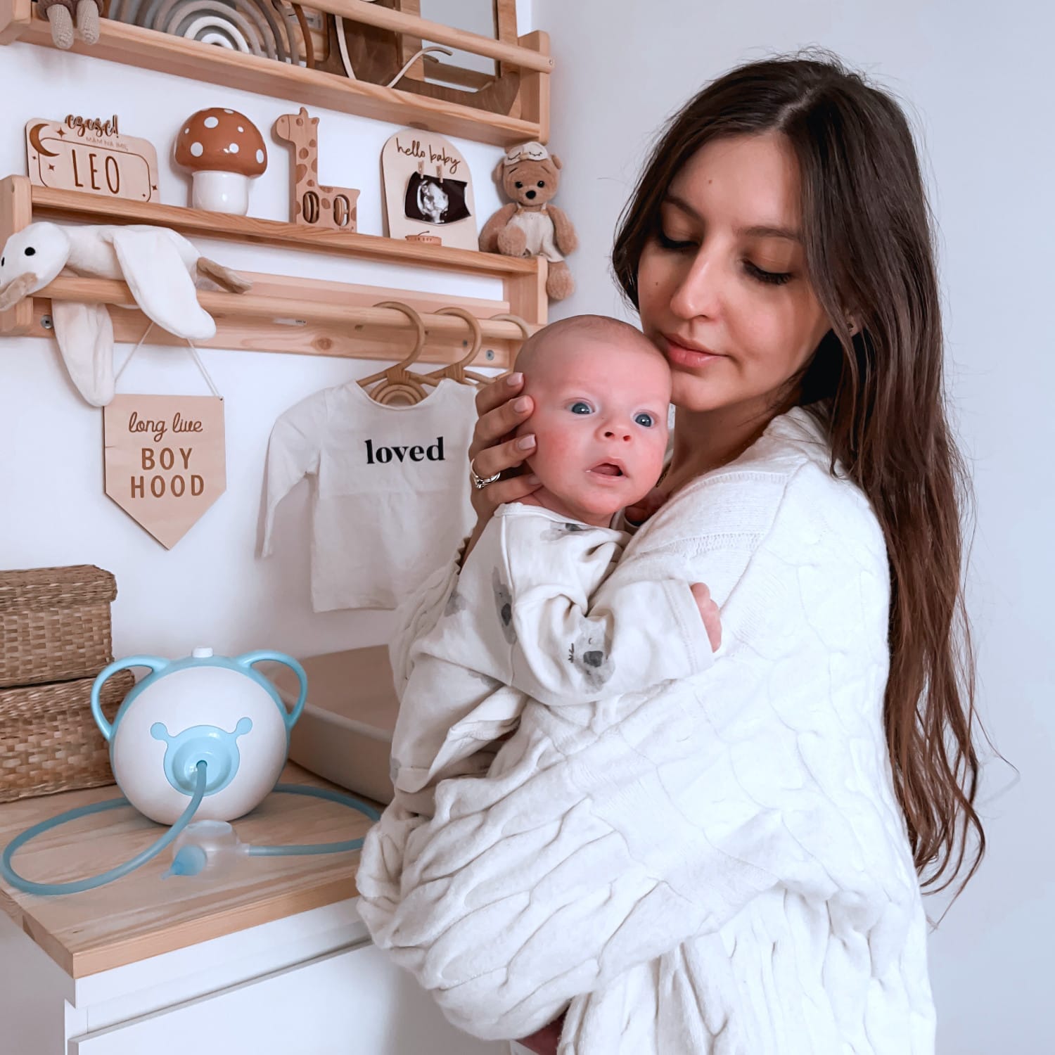 Une mère tenant son joli nouveau-né dans les bras, debout dans une chambre d'enfant à côté d'un mouche-bébé électrique Nosiboo Pro.