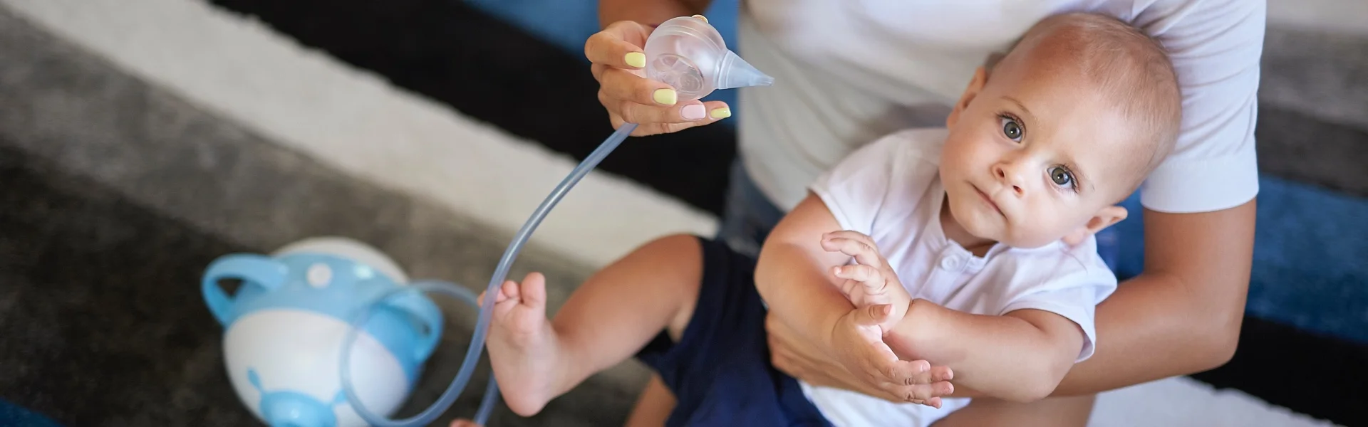 Un petit garçon regarde dans la caméra, tenu par sa mère qui va lui dégager le nez à l'aide du mouche-bébé électrique Nosiboo Pro