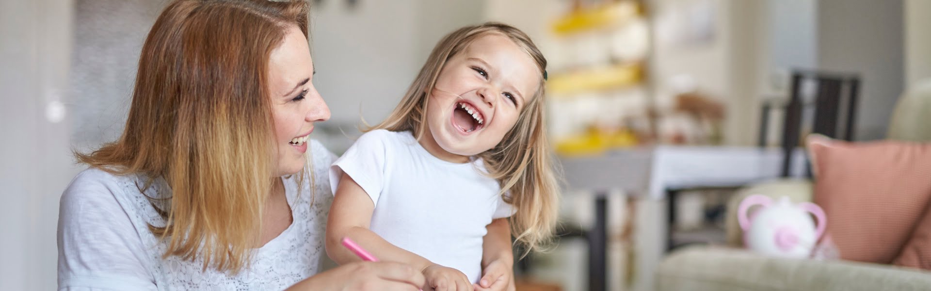 Une mère et sa petite fille dessinant et riant ensemble, avec un mouche-bébé électrique Nosiboo Pro rose sur le canapé en arrière-plan