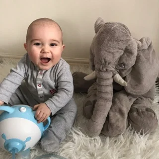 Un bébé amusé assis sur un tapis à côté d'un éléphant en peluche, tenant la tête de nounours du mouche-bébé électrique Nosiboo Pro bleu par les oreilles