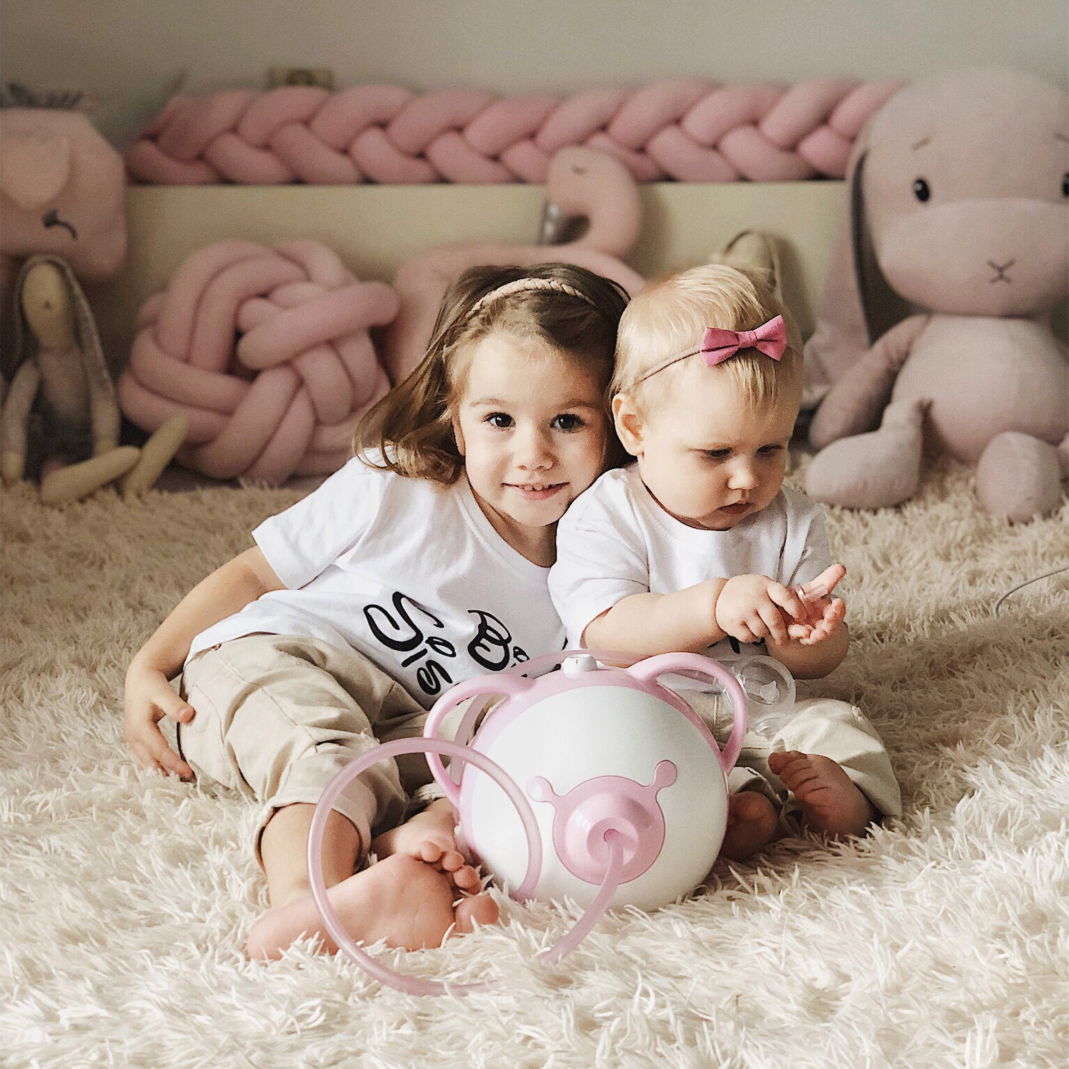 Deux jolies petites filles assises l'une à côté de l'autre sur un lit avec un mouche-bébé électrique Nosiboo Pro rose