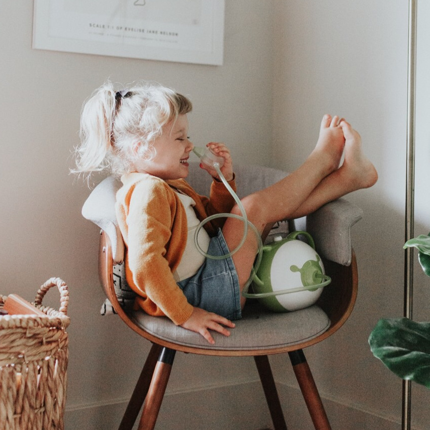 Une petite fille heureuse assise sur une chaise avec un mouche-bébé électrique Nosiboo Pro vert