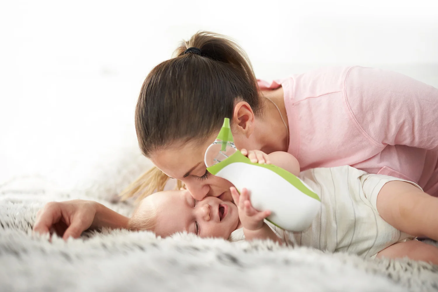 Une mère embrasse son bébé qui tient un mouche-bébé portatif Nosiboo Go dans ses mains, allongé sur un lit