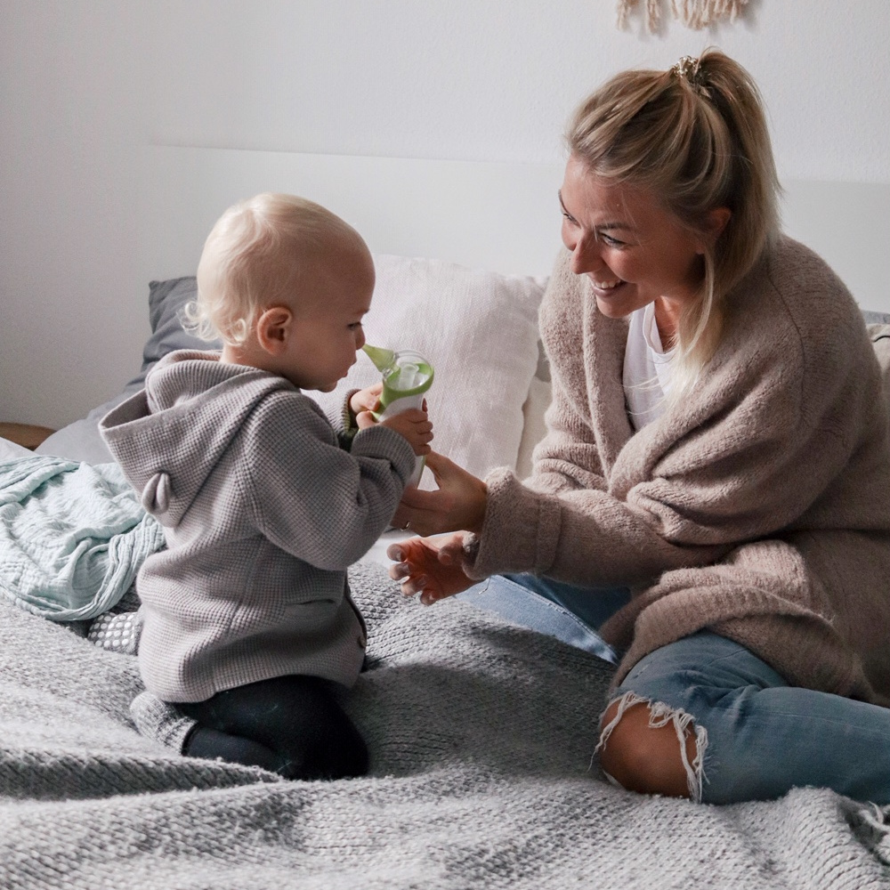 Une mère et son fils, assis sur le lit, aspirent le nez de l'enfant avec le mouche-bébé électrique portatif Nosiboo Go