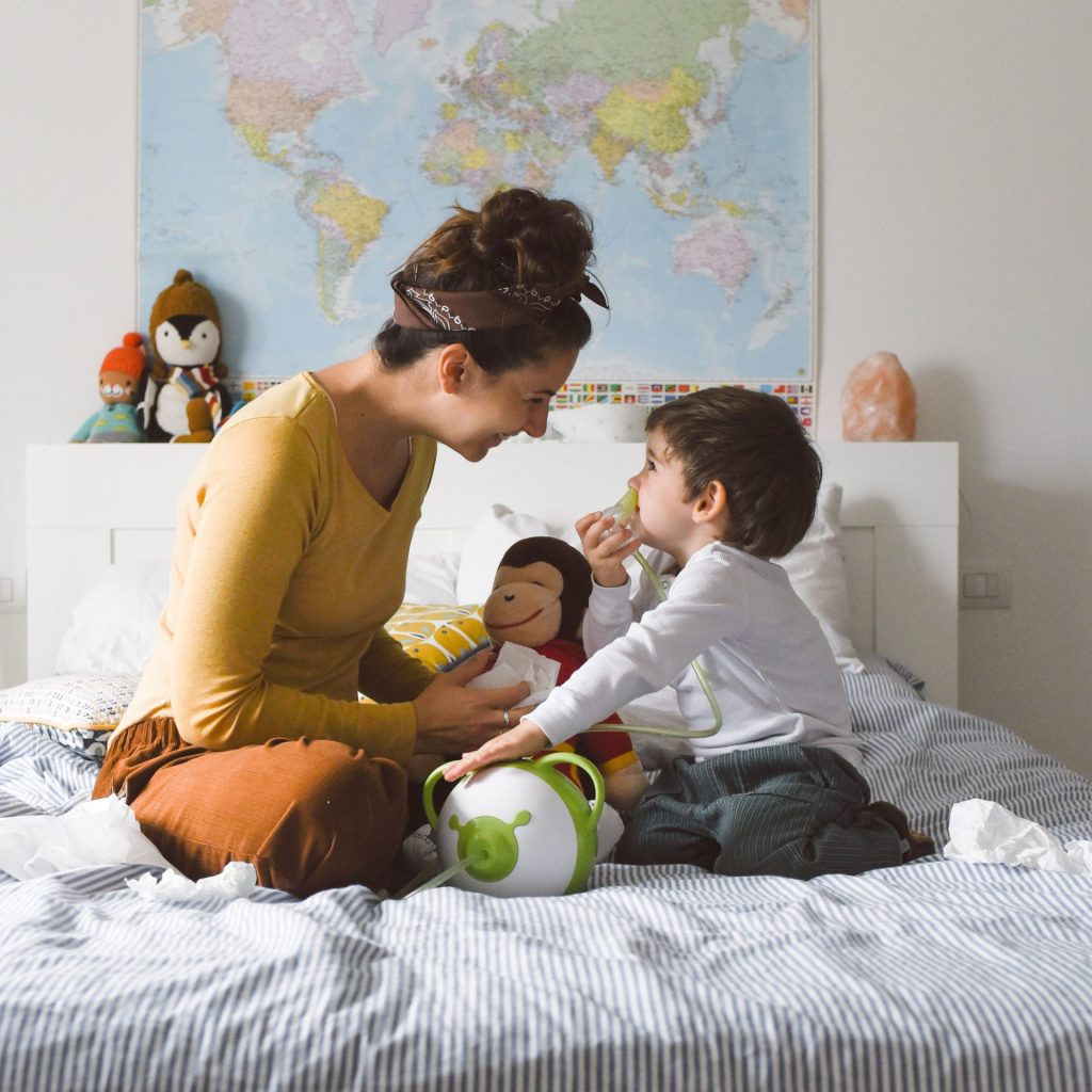 Une mère et son fils, assis sur le lit, aspirent le nez de l'enfant avec le mouche-bébé électrique Nosiboo Pro