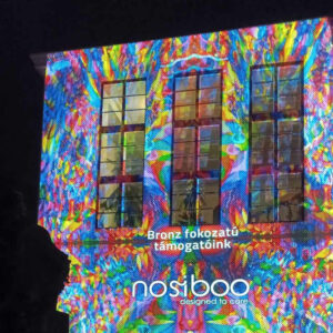 Le logo de Nosiboo s'affiche avec une lumière colorée sur un bâtiment, informant du niveau bronze du parrainage du Festival des Lumières de Zsolnay à Pécs.