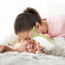 Une mère embrasse son bébé qui tient un mouche-bébé portatif Nosiboo Go dans ses mains, allongé sur un lit